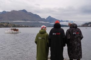 Three swimmers wearing dryrobes getting ready to do open water swimming in Lake Whakatipu, Queenstown on a moody, grey day.