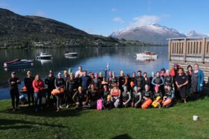 A group of more than 40 swimmers in wetsuits stands beside lake Wahakatipu with mountains in the background