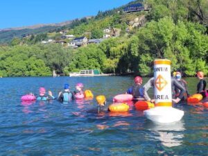 A white top hat buoy floats in lake Whakatipu and is surrounded by swimmers wearing colourful tow floats